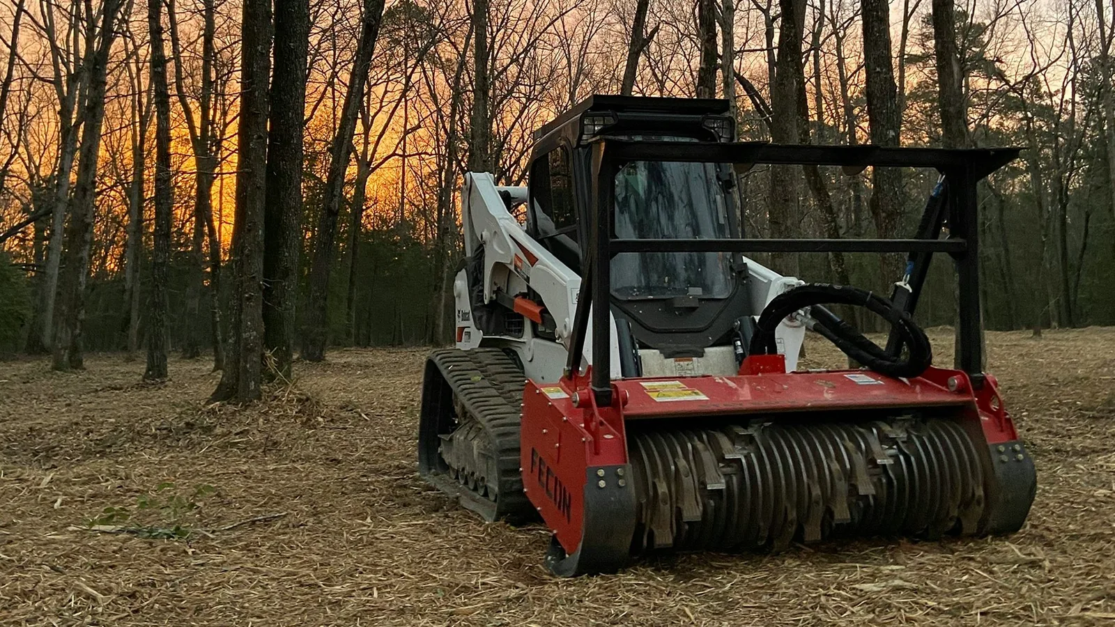 Compact track loader with a drum mulcher head parked on a mulched clearing as the sun sets behind the timber