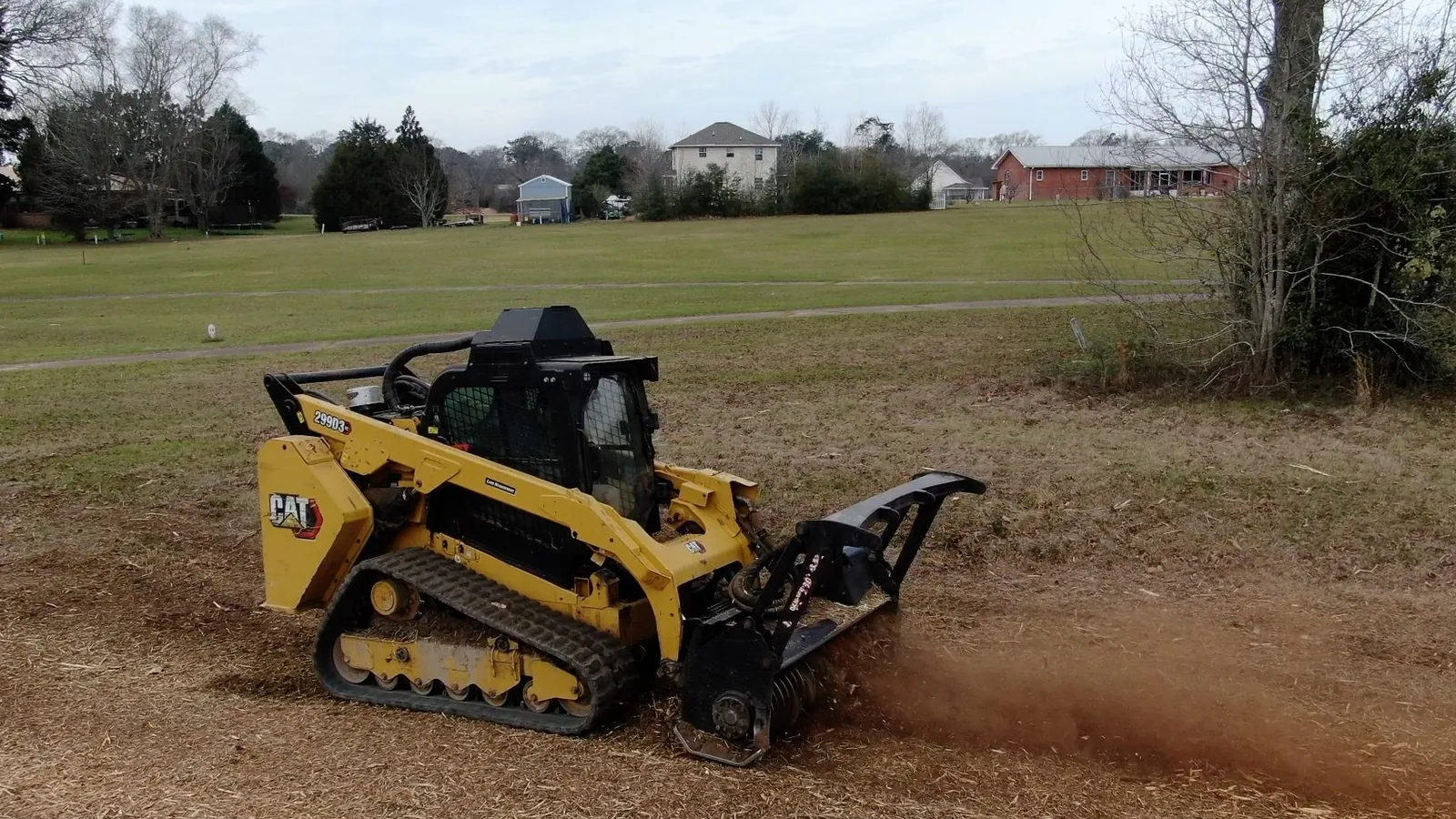 Compact track loader running a mulcher head through standing brush, throwing a cloud of mulch