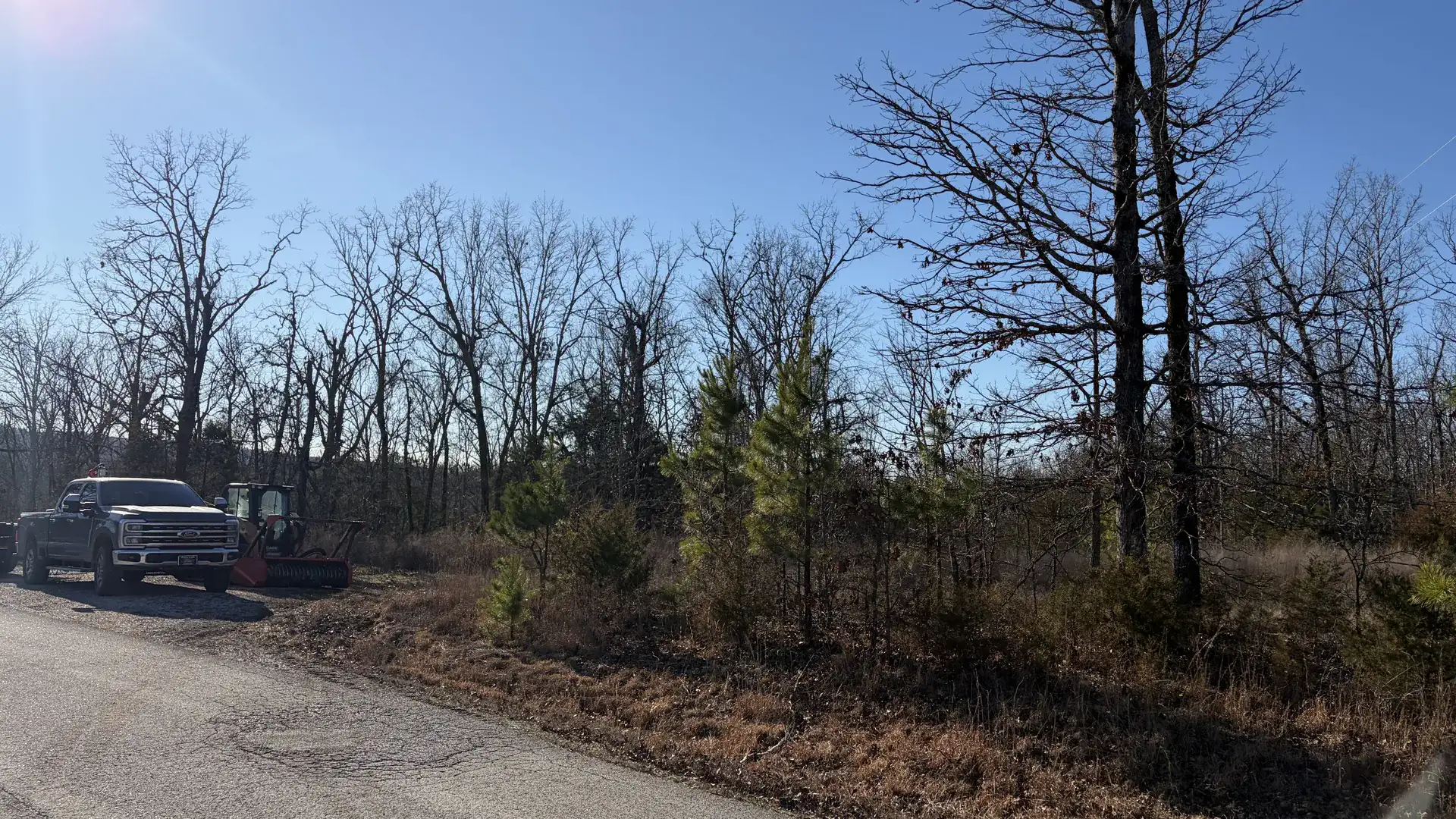 Wooded roadside lot with a pickup and a compact track loader parked at the edge of the timber in Northeast Arkansas