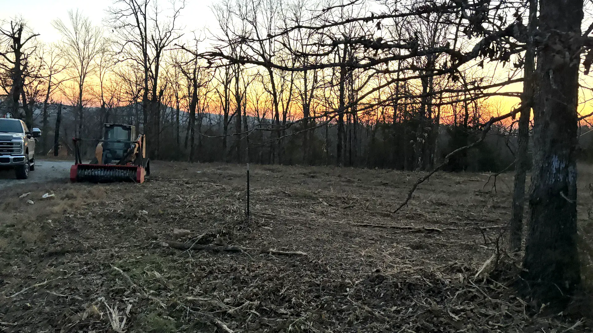 Cleared and mulched lot at sunset with a track loader and a pickup parked at the treeline in Northeast Arkansas