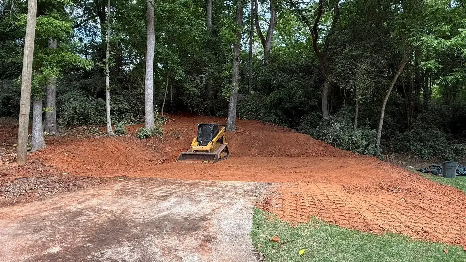 Compact track loader rough-grading a freshly cleared red-clay lot at the edge of the woods